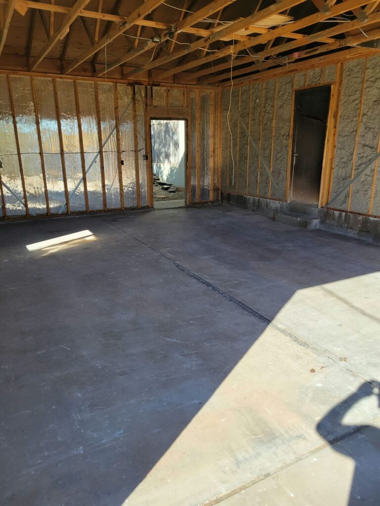 Interior of a partially demolished building with exposed wooden framing, reflective insulation on some walls, and a concrete floor. The ceiling shows wooden beams with visible wiring, and there are two doorways leading to other areas. The image highlights a demolition project by Cutting Edge Demolition.