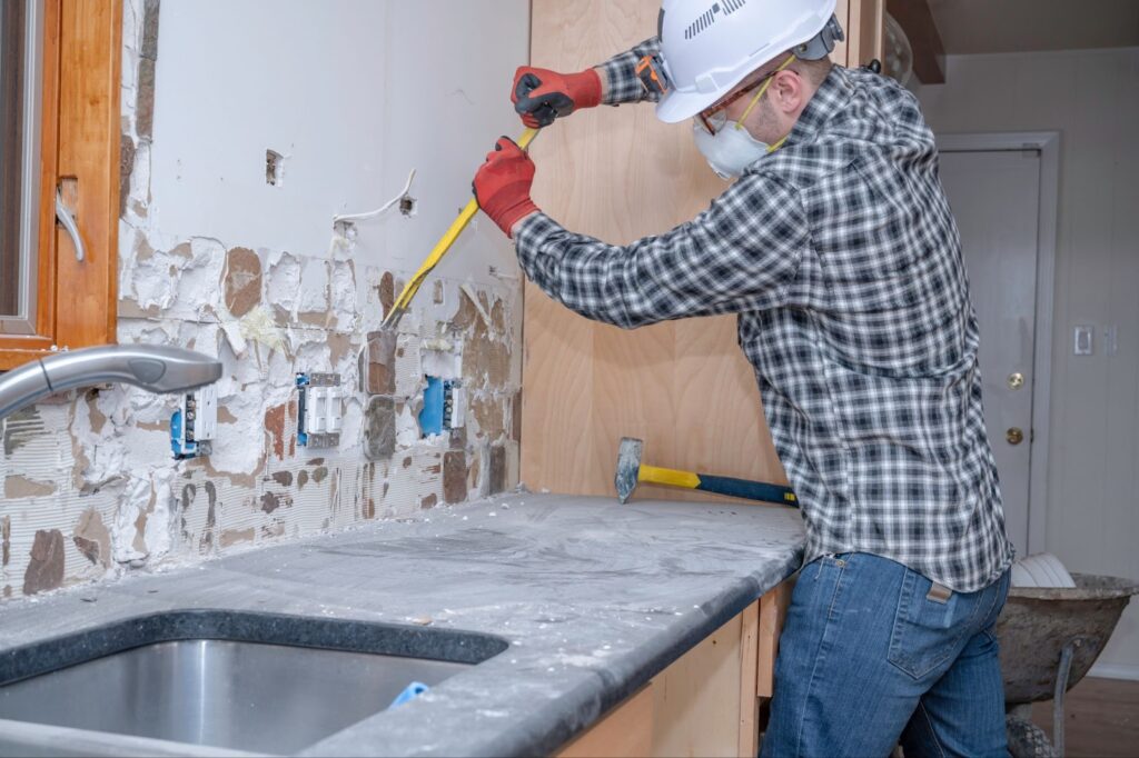 A professional worker in a hard hat and safety glasses is demolishing a kitchen counter
