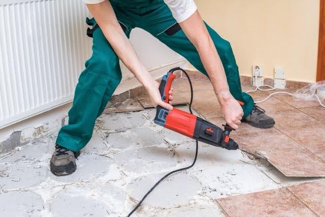 A man in green overalls repairs a tile floor using a power tool focused on his task in a well-lit room