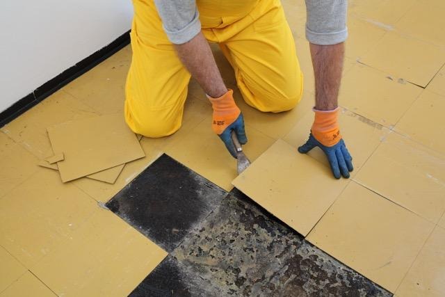 A person wearing yellow pants and orange gloves uses a tool to remove yellow floor tiles