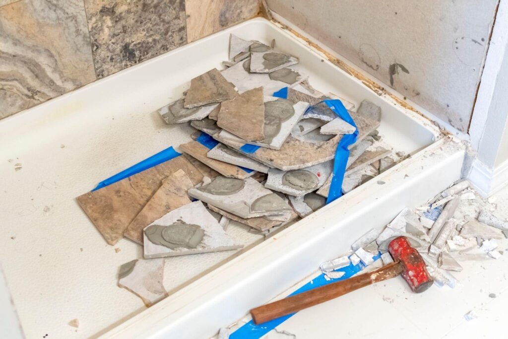 A pile of broken tiles with adhesive rests in a white shower base surrounded by debris