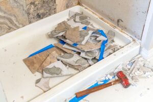 A pile of broken tiles with adhesive rests in a white shower base surrounded by debris