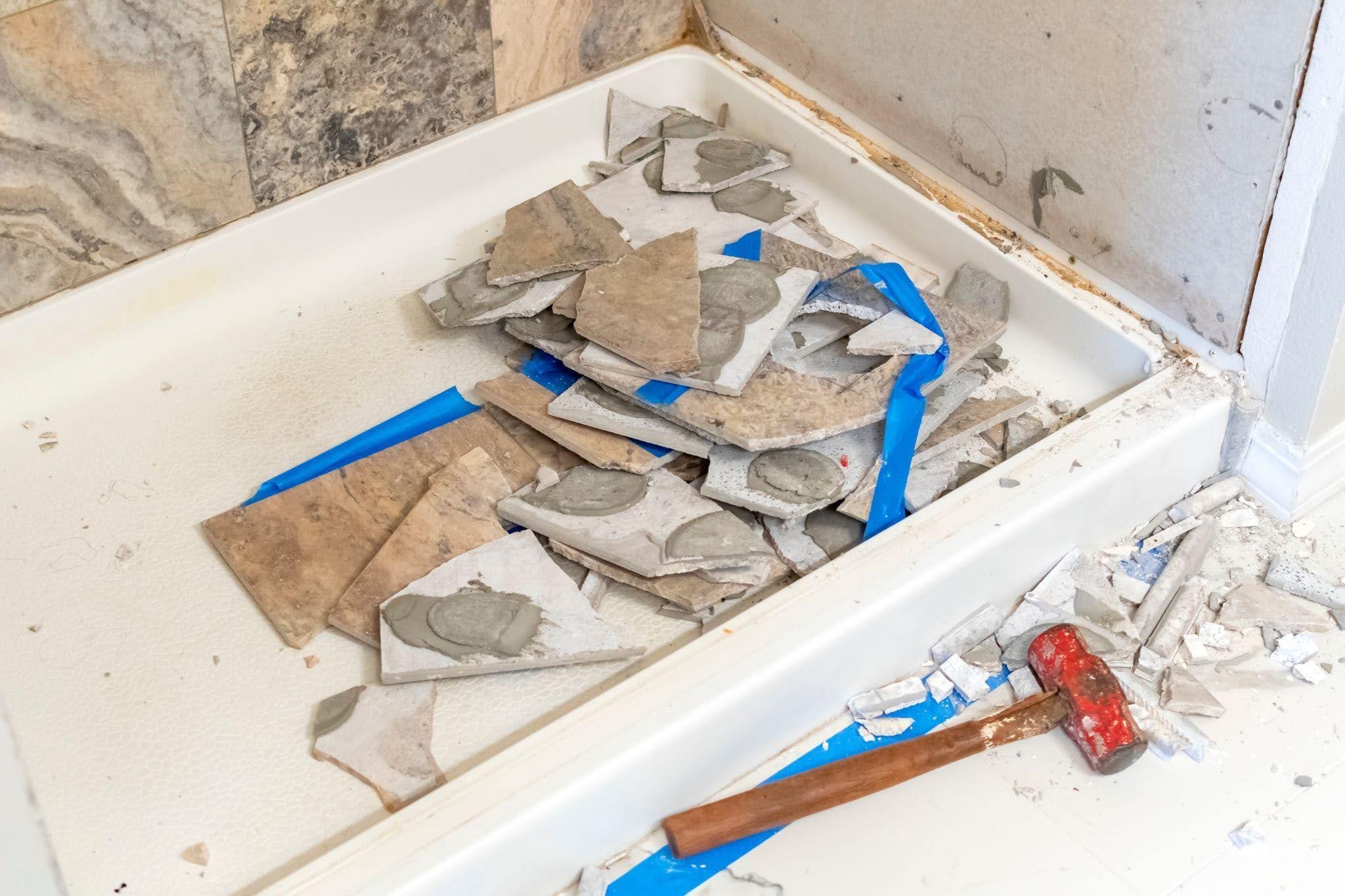 A pile of broken tiles with adhesive rests in a white shower base surrounded by debris