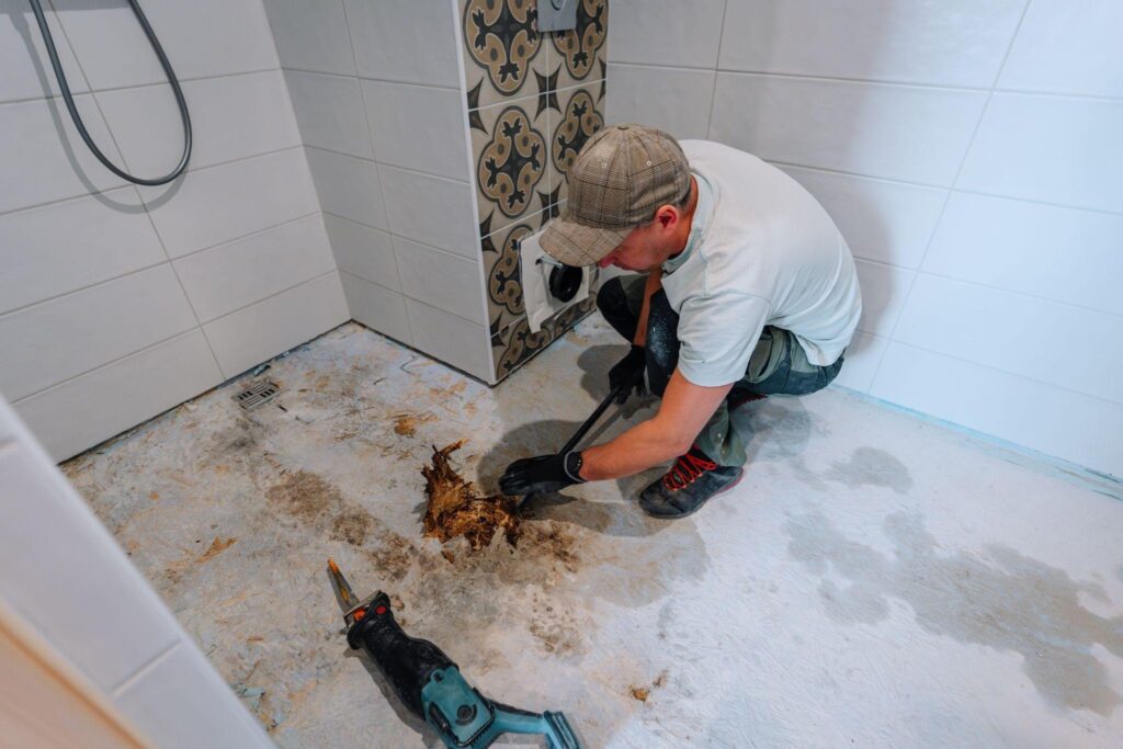 A man wearing a cap and gloves crouches on a bathroom floor using a tool to remove damaged material