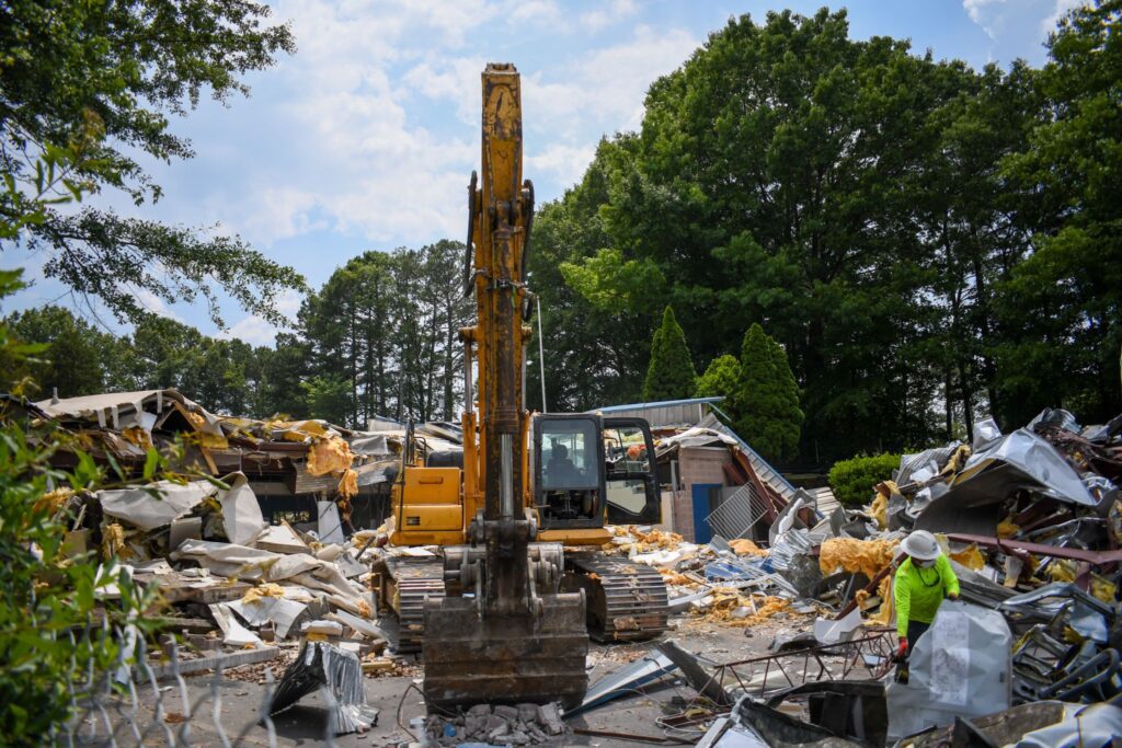 An excavator at a construction site demolishing a building surrounded by debris and trees.