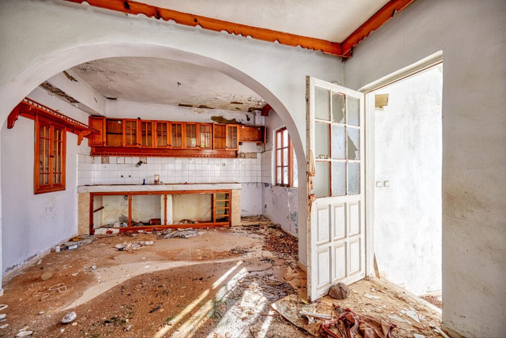 Abandoned, dilapidated kitchen with peeling paint, debris on the floor, and old wooden cabinetry framed by a large archway.