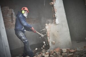 Construction worker wearing safety gear using a sledgehammer to demolish a interior brick wall.