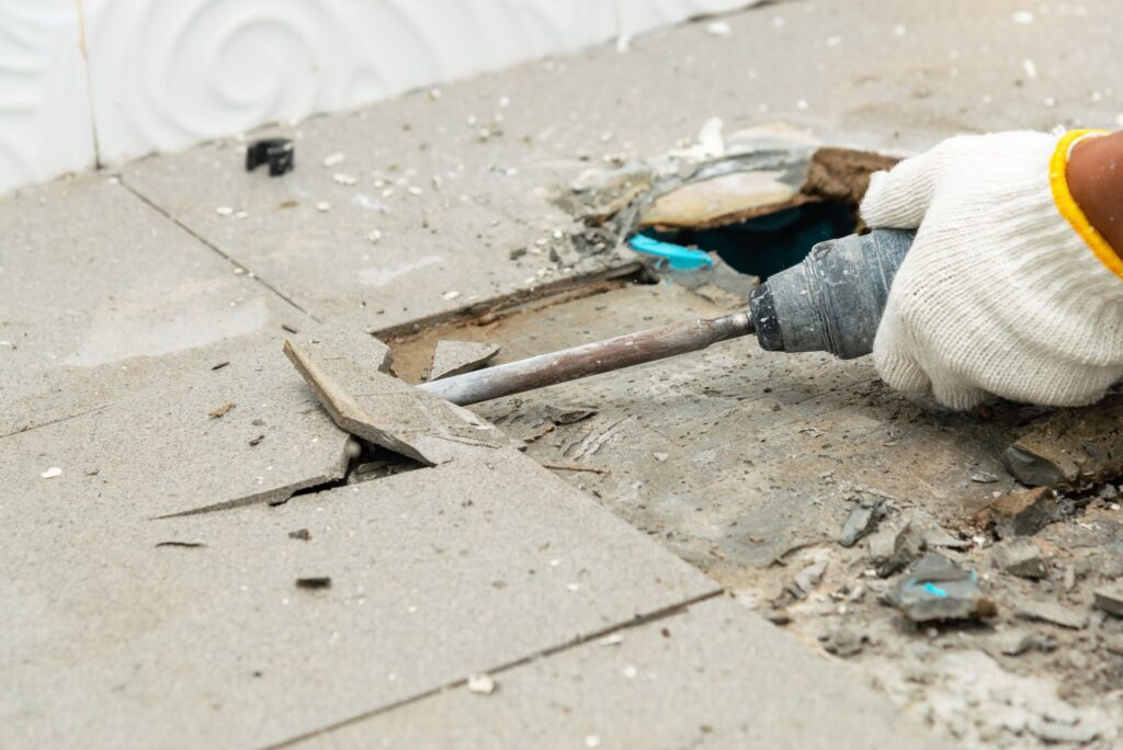 Gloved hand using a chisel attachment on a power tool to remove broken grey floor tiles.